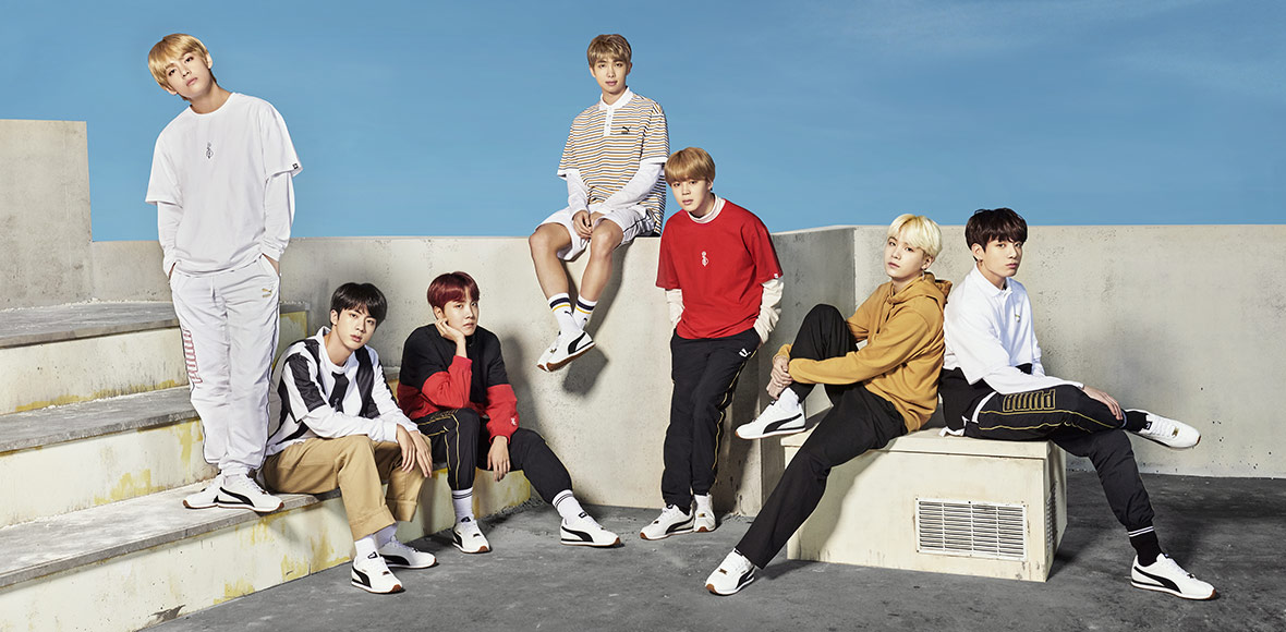 Members of BTS posed on white steps and partitions against a blue sky, wearing clothes from their line. 