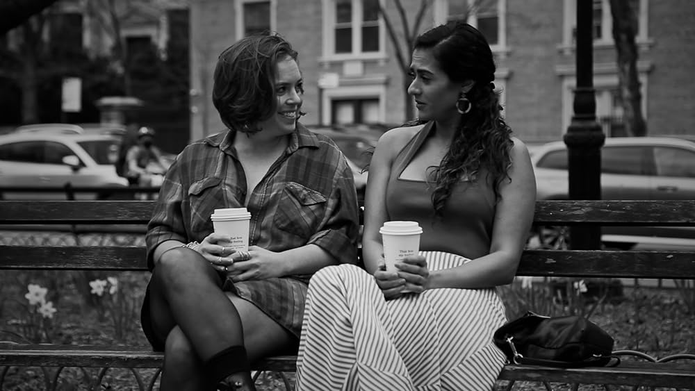 Katie Muldowney and Kavita Jariwala are sitting on a park bench conversing as they hold takeout coffee cups. 